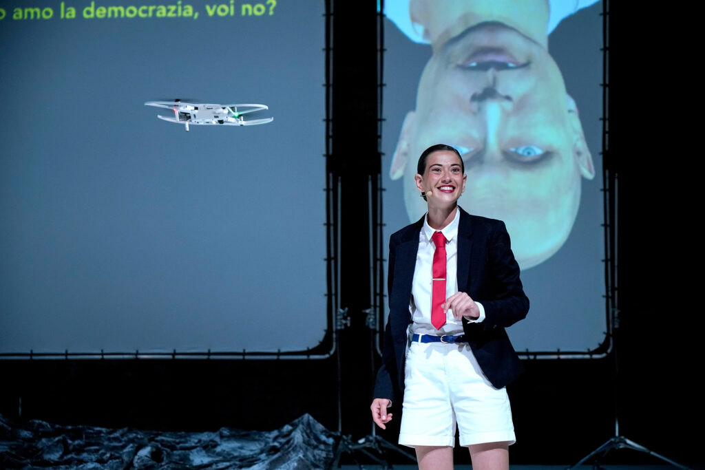 L'actrice Anna Pérez en veste de costume noire, short blanc et cravate rouge, devant une projection du visage de Poutine à l'envers, dans le spectacle The Mountain, au Paris Globe Festival.