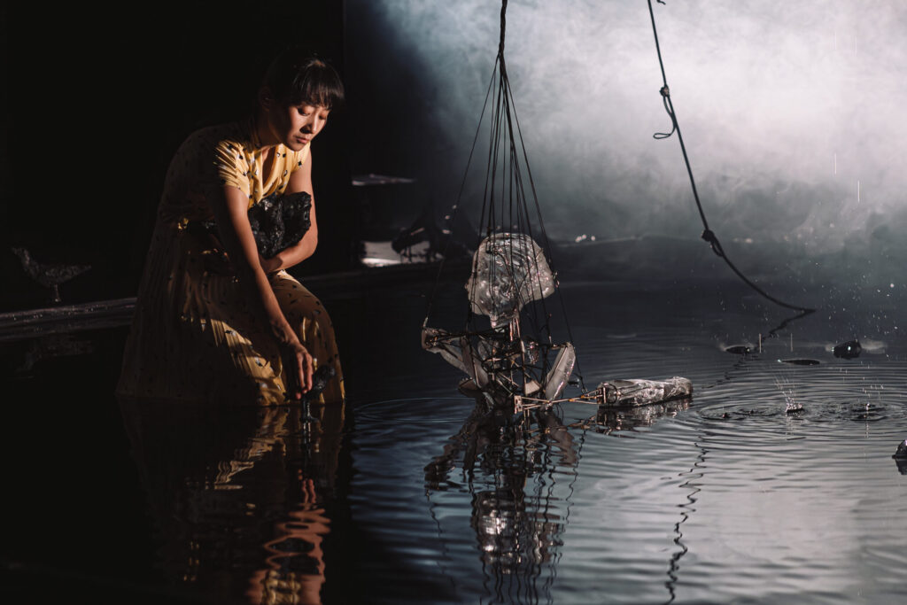 Une comédienne du spectacle Les Vagues, d’Élise Vigneron, agenouillée près d'une marionnette de glace finissant de fondre au milieu d'une flaque d'eau.