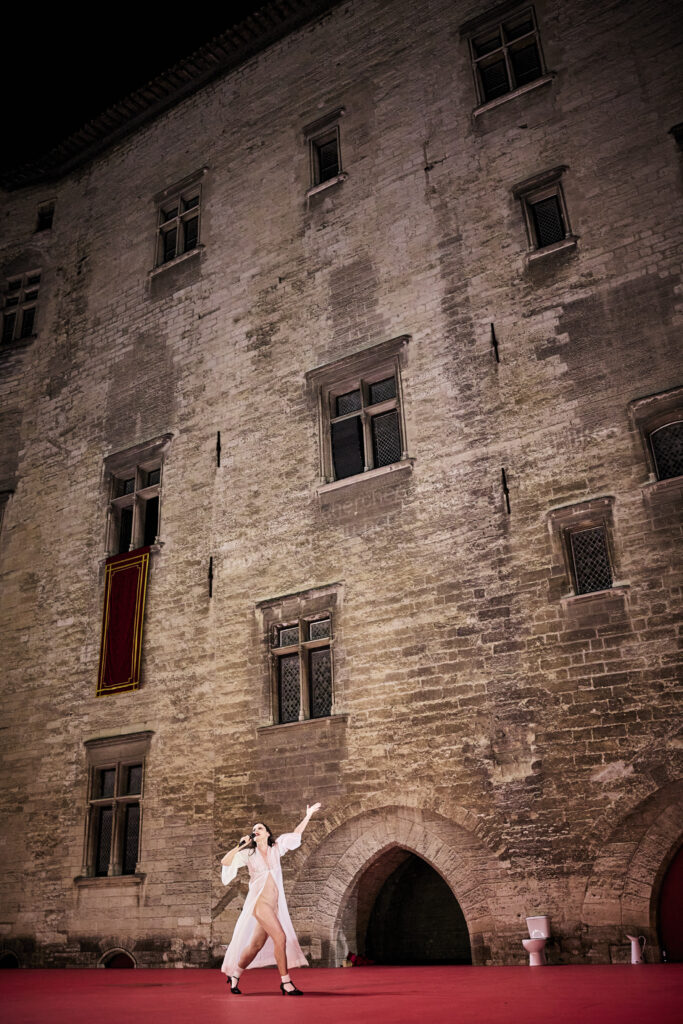 Angelica Liddell nue dans un déshabillé blanc, micro à la main, dans la cour d'Honneur du Palais des Papes, dans le spectacle DÄMON, au festival d'Avignon 2024