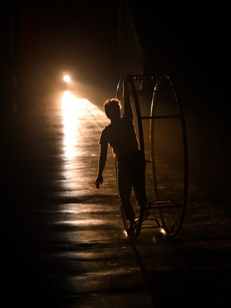 L'artiste circassien Noa Aubry et sa roue allemande, éclairés à contrejour par un projecteur, dans le spectacle "Là où la nuit n'est pas si loin puisque le jour s'en va déjà", lors du festival révélations circusnext au Théâtre de la cité internationale.